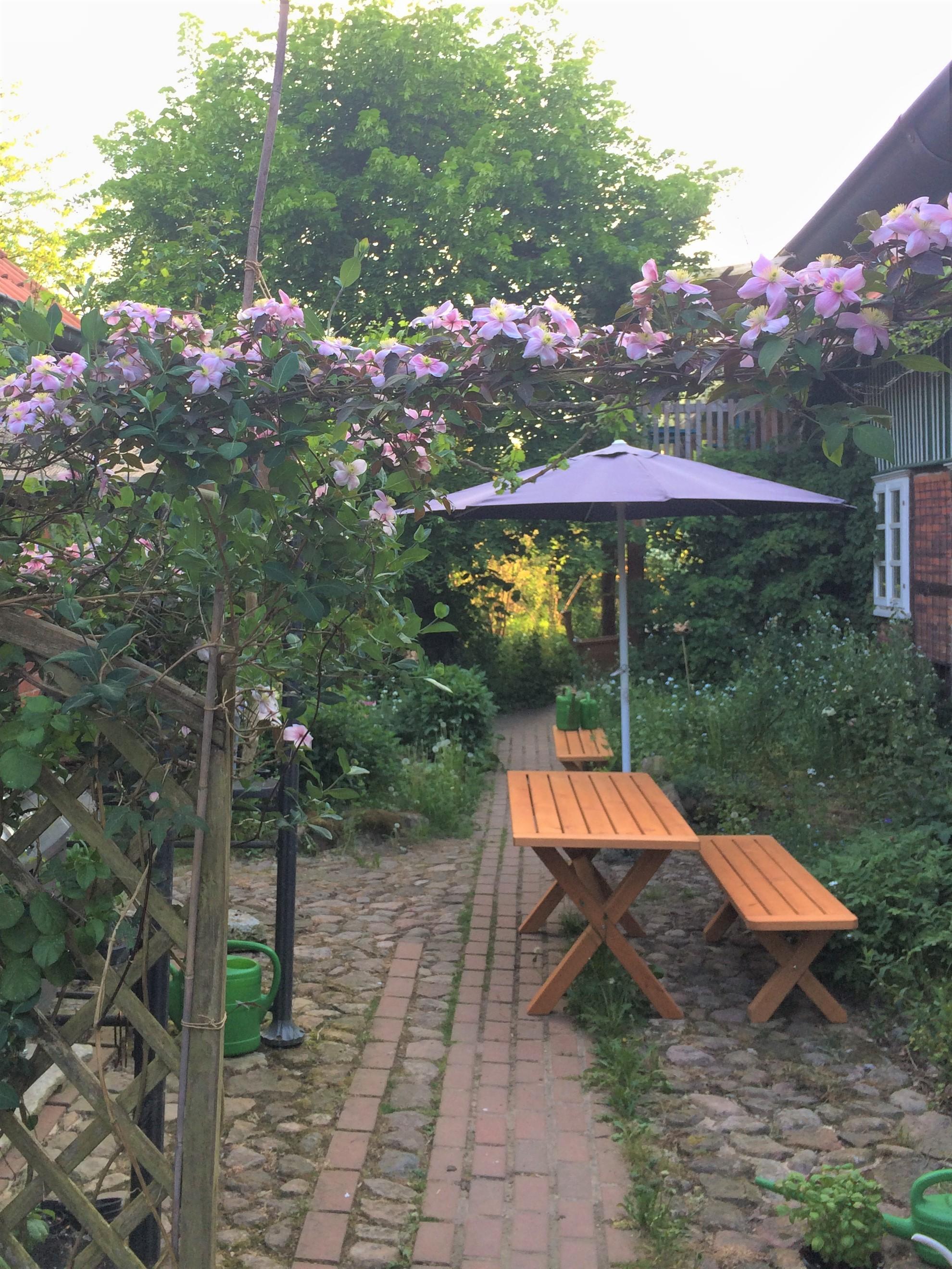 das Bild zeigt einen Sitzplatz im Privelacker Paradiesgarten, unter einem Sonnenschirm,umrahmt mit Clematisblüten.hmt mit Privelacker Paradiesgarten