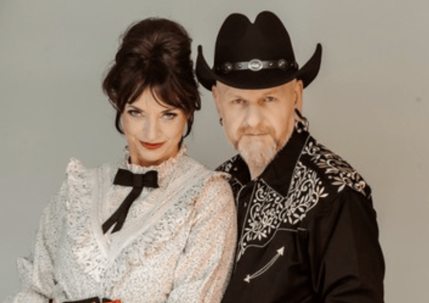 Couple in Western costumes: woman in a lace white dress with a black bow beside a man in a black embroidered shirt and cowboy hat.