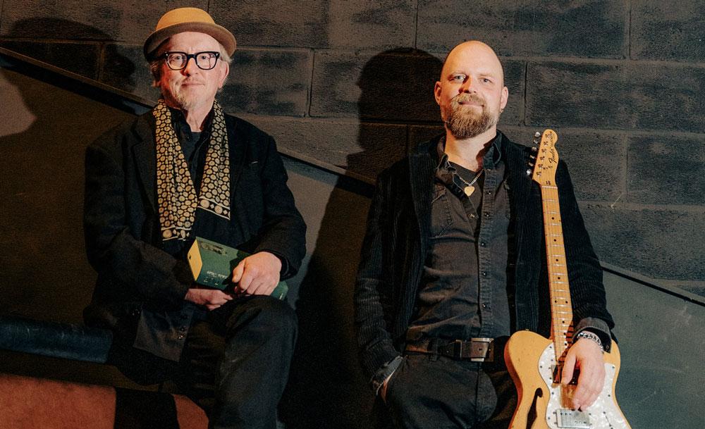 Two men pose in a dim stairwell; one wears a hat and glasses and holds a small green booklet, the other holds a yellow guitar in front of a brick wall.