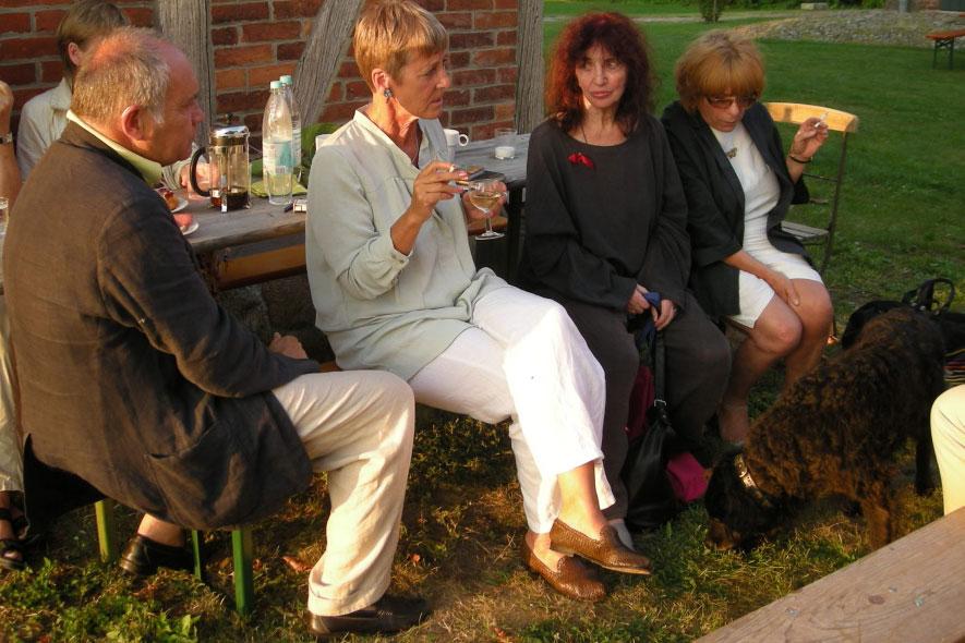 Group of five adults seated at a picnic table outdoors, chatting with drinks nearby and a dog at their feet.