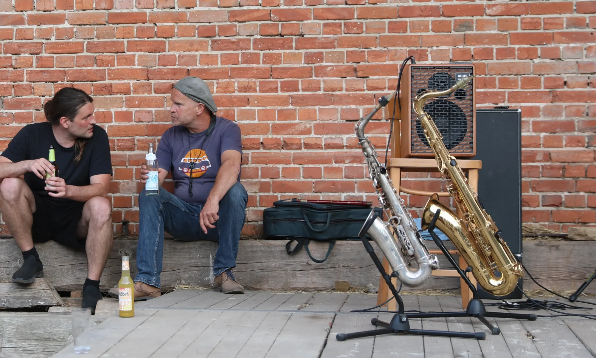 Two men sit on a wooden platform against a red brick wall, each holding a bottle and talking, with brass instruments and an amp nearby.