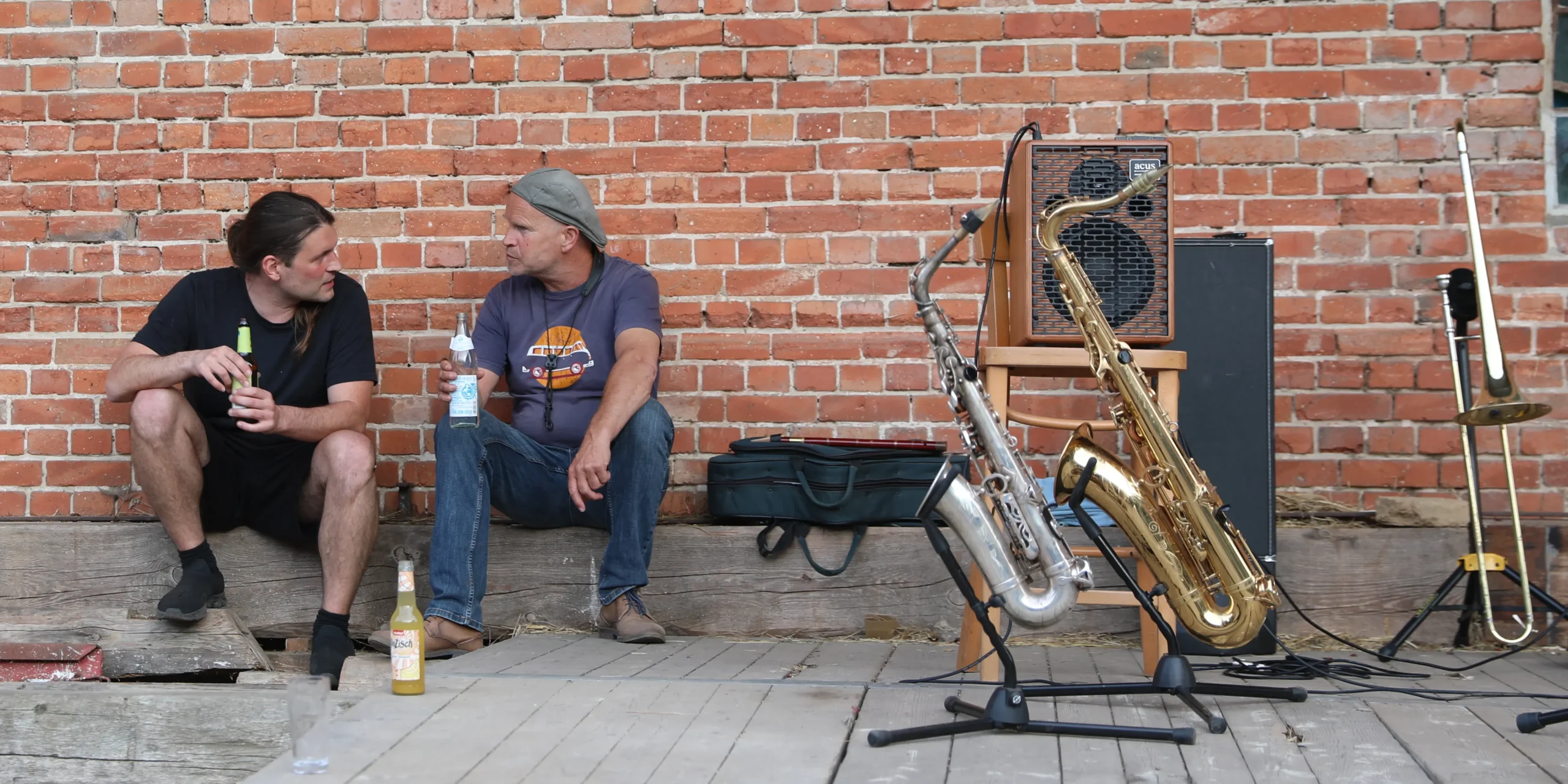 Two men sit on a wooden platform against a red brick wall, each holding a bottle and talking, with brass instruments and an amp nearby.